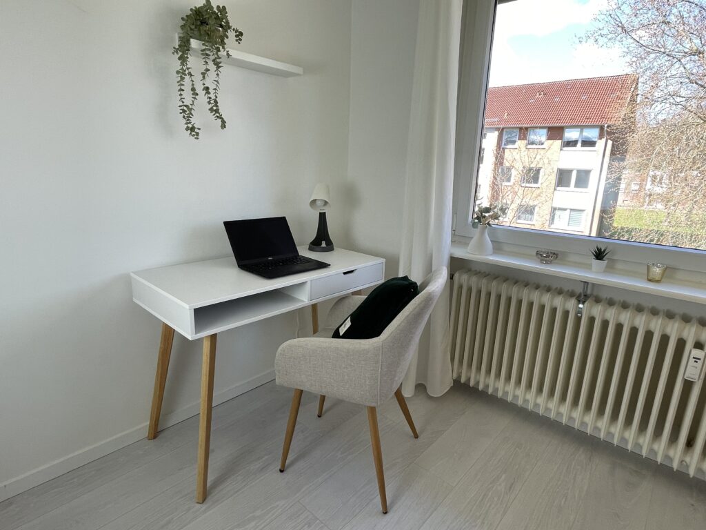 Nahaufnahme Arbeitsecke am Fenster Zeigt: Schreibtisch, Laptop, Fensterblick, Tischlampe, Stuhl, Regalbrett mit Hängepfalnze